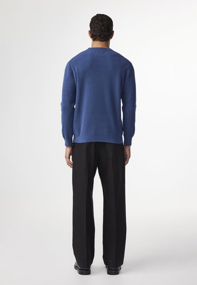Maglione a maglia blu con texture, colletto rotondo e dettagli a coste sui polsini, abbinato a pantaloni neri a gamba larga, mostrando una vista posteriore casual.