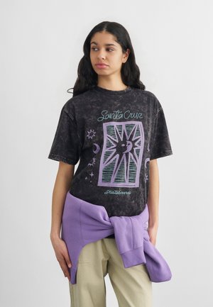 Noor naine, pikkade tumedate juustega, kes kannab musta "Santa Cruz" graafilise kujutisega t-särki, beeže pükse ja tal on vöökoha ümber seotud lilla dressipluus.
