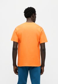 Πορτοκαλί βαμβακερό μπλουζάκι με κοντά μανίκια, στρογγυλή λαιμόκοψη και άνετη εφαρμογή· φοριέται με μπλε παντελόνια, από πίσω όψη.