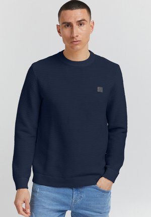 Pulover albastru marin, cu nervuri, cu mâneci lungi, guler rotund și un mic logo brodat pe piept. Tesătura prezintă o textură și un model subtil.