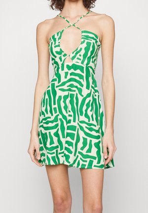 Mujer vistiendo un mini vestido sin mangas con patrón verde y blanco, con tirantes halter que se cruzan en el pecho y un corte en forma de ojo de cerradura en la parte frontal.