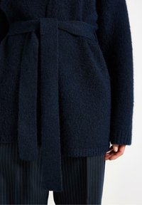Cardigan a maglia testurizzata blu navy con cintura, dotato di maniche lunghe e polsini a coste. Abbinato a pantaloni a righe.