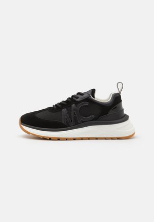 Zwarte sneaker met mesh en suède, dikke witte zool, gumkleurige buitenzool, MC-logo aan de zijkant, vetersluiting en trekbandje bij de hiel.