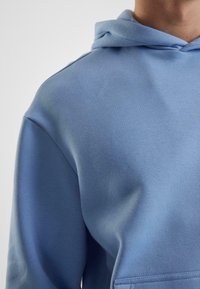 Lyseblå hettegenser laget av mykt stoff. Har en hette med snor og en frontlomme med pene sømmer. Glatt tekstur og uformelt design.