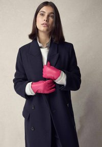 Pink leather gloves worn by a person in a dark navy coat, layered over a white sweater with a striped collar. Smooth texture, fitted design.