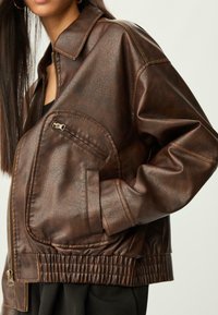Femme portant une veste en cuir brun foncé texturée avec une fermeture éclair et une poche zippée, la main dans la poche de la veste, sur un fond uni.