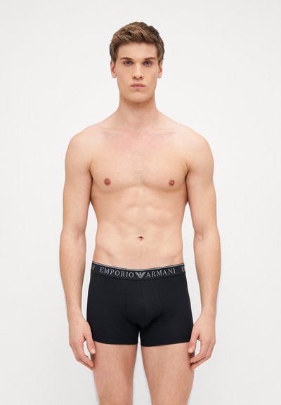 Čierne boxerky s elastickým pásom s nápisom "EMPORIO ARMANI" v bielej farbe. Hladká tkanina a priliehavý strih, zobrazené na holom trupe.