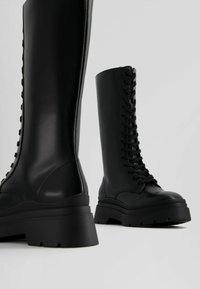 Bottes noires en cuir montant jusqu'au genou avec un laçage à l'avant, semelle épaisse en caoutchouc et texture lisse, dotées d'un bout arrondi et d'un design minimaliste.