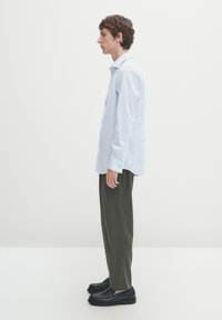 Helleblauses Langarmshirt mit Kragen, kombiniert mit olivgrünen Hosen und schwarzen Schuhen. Glatte Stoffqualität, taillierte Passform und Knopfdetails.