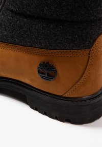 Botte en cuir marron avec tige en feutre noir, présentant un logo noir et une semelle noire robuste. Coutures visibles le long des coutures.