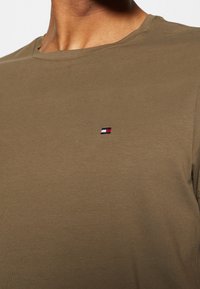 Oliivinvihreä puuvillainen t-paita, jossa on pyöreä pääntie; rinnassa pieni kirjailtu logo punaisen, valkoisen ja sinisen sävyissä.