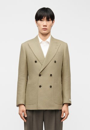 Jonge man draagt een beige dubbelbreasted blazer over een wit overhemd en grijze broek, staand tegen een effen lichte achtergrond.