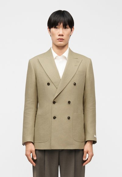 Joven con blazer cruzado beige sobre una camisa blanca y pantalones grises, posando contra un fondo claro y sencillo.