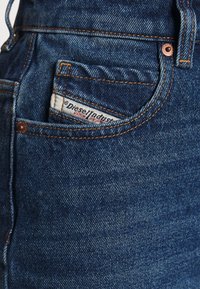 Jeans de mezclilla azul oscuro que cuentan con un pequeño bolsillo frontal con una etiqueta de marca visible. Costuras y herrajes en tonos bronce.