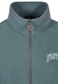 Fleece-takki turkoosina, jossa on pystytakki, täysi vetoketju ja valkoinen "STARTER"-logo vasemmalla puolella. Teksturoitu kangas, jossa on hienovarainen kiilto.