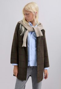 Mujer rubia que lleva un abrigo de cuadros marrón sobre una camisa azul, con un suéter beige drapeado y atado alrededor de los hombros.