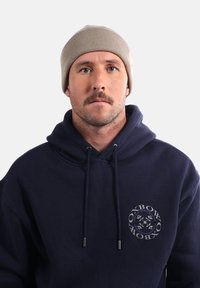 Bonnet en tricot côtelé beige et hoodie bleu foncé avec logo. Le hoodie est doté de cordons de serrage et d'un design circulaire sur le devant.