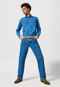 Ung mand stående med brede ben i blå denimskjorte, jeans, lys bælt og brune støvler mod en ensfarvet lys baggrund.