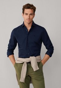 Camisa azul marino de botones con mangas largas, de suave tela de algodón, combinada con pantalones verde oliva y un suéter de punto beige atado a la cintura.
