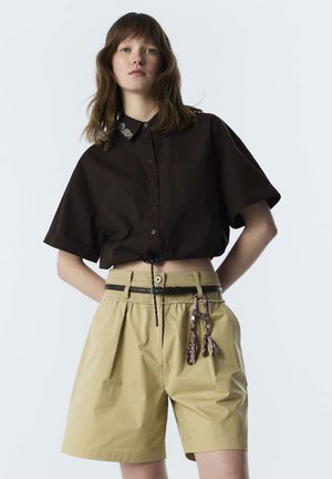 Junge Frau trägt ein locker sitzendes dunkelbraunes, geknöpftes Hemd und beige, hoch taillierte Shorts mit einem schwarzen Gürtel und einem gemusterten Schlüsselanhänger.
