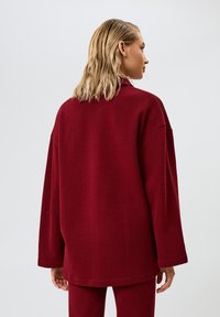 Camicia rossa a costine oversize con colletto alto, spalle scese e maniche lunghe. Tessuto morbido con vestibilità rilassata, che termina a metà fianchi.