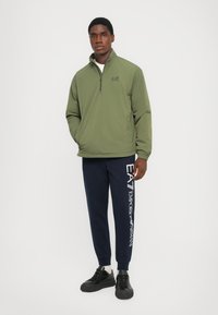 Oliven grønn kvart-zip pullover med ståkrage; kombinert med marineblå joggebukser med hvit logotrykk; svarte joggesko.
