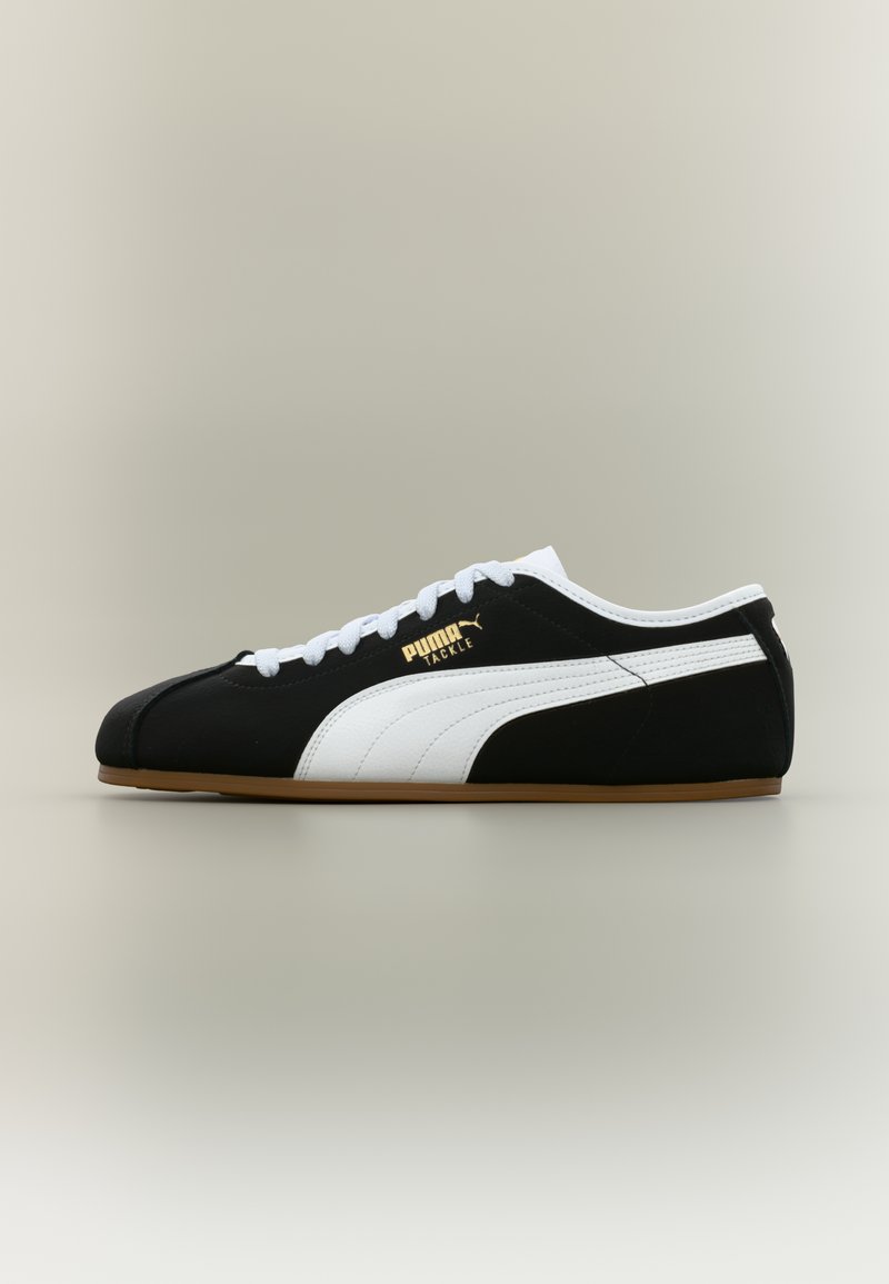 Μαύρο αθλητικό παπούτσι Puma Tackle με λευκή ρίγα, λευκά κορδόνια και σανδαλί ταμπά, εμφανισμένο σε πλευρική όψη αριστερά σε ουδέτερο φόντο.