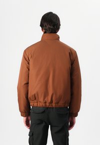 Brun polstret jakke med høj krave, elastiske ærmer og talje. Glat tekstur og simpelt design; båret med mørke cargo bukser.