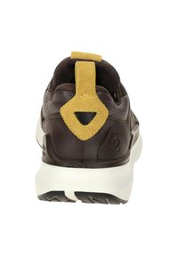 Zapato deportivo marrón con un collar acolchado, una lengüeta de ante amarilla y un upper texturizado. Presenta una media suela blanca y una suela con patrón.