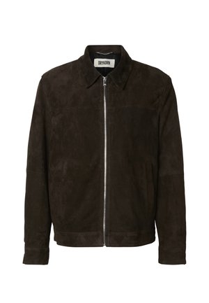 Jeune homme debout, les mains dans les poches, portant une veste zippée marron foncé, un pantalon beige à carreaux et des bottes en cuir noires.