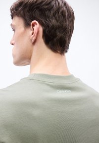 Νέος άνδρας με κοντά καστανά μαλλιά φορώντας ένα ανοιχτοπράσινο t-shirt Calvin Klein, που απεικονίζεται από πίσω πάνω σε απλό λευκό φόντο.