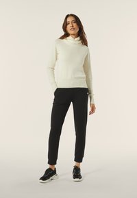 Maglione beige chiaro con scollo a costine, abbinato a pantaloni neri a sigaretta con tasche laterali e un sottile dettaglio in metallo, e sneakers nere.