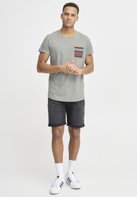 Grå kortermet t-skjorte med mønstret lomme, kombinert med mørk denimshorts og hvite joggesko. Enkel, uformell design.