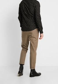 Camicia nera con piccoli pois dorati, abbinata a pantaloni slim-fit marrone chiaro e stivaletti neri. Tessuto liscio e taglio ordinato.