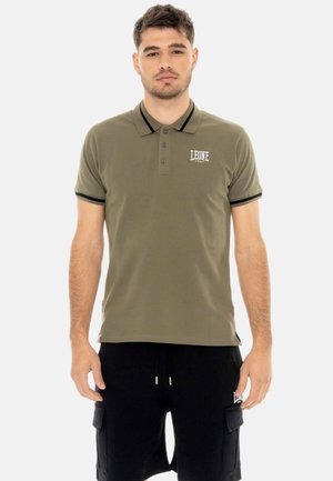 Man draagt een olijfgroen poloshirt met zwarte biezen en zwarte cargoshorts, staand tegen een effen lichte achtergrond.