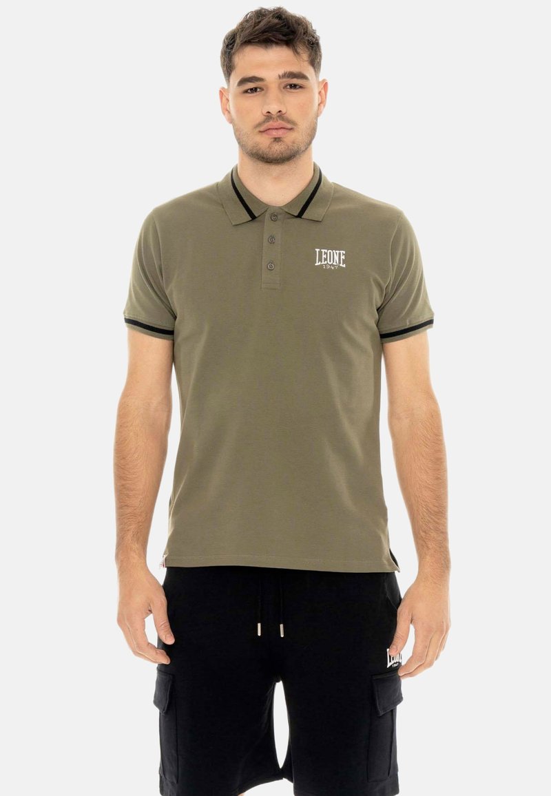 Homme portant un polo vert olive avec des finitions noires et un short cargo noir, debout devant un fond clair uni.