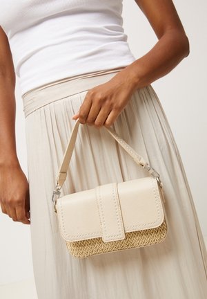 Femme portant un débardeur blanc et une jupe beige tenant un petit sac à main beige avec un rabat en cuir et un fond tissé.