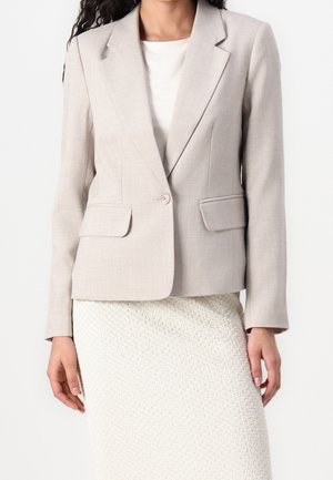 Mujer con blazer beige claro de un botón sobre una blusa blanca y falda blanca texturizada, posando frente a un fondo liso.