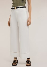 White wide-leg trousers feature a smooth texture with cut-out detailing at the hem. Paired with a black belt and strappy black sandals.