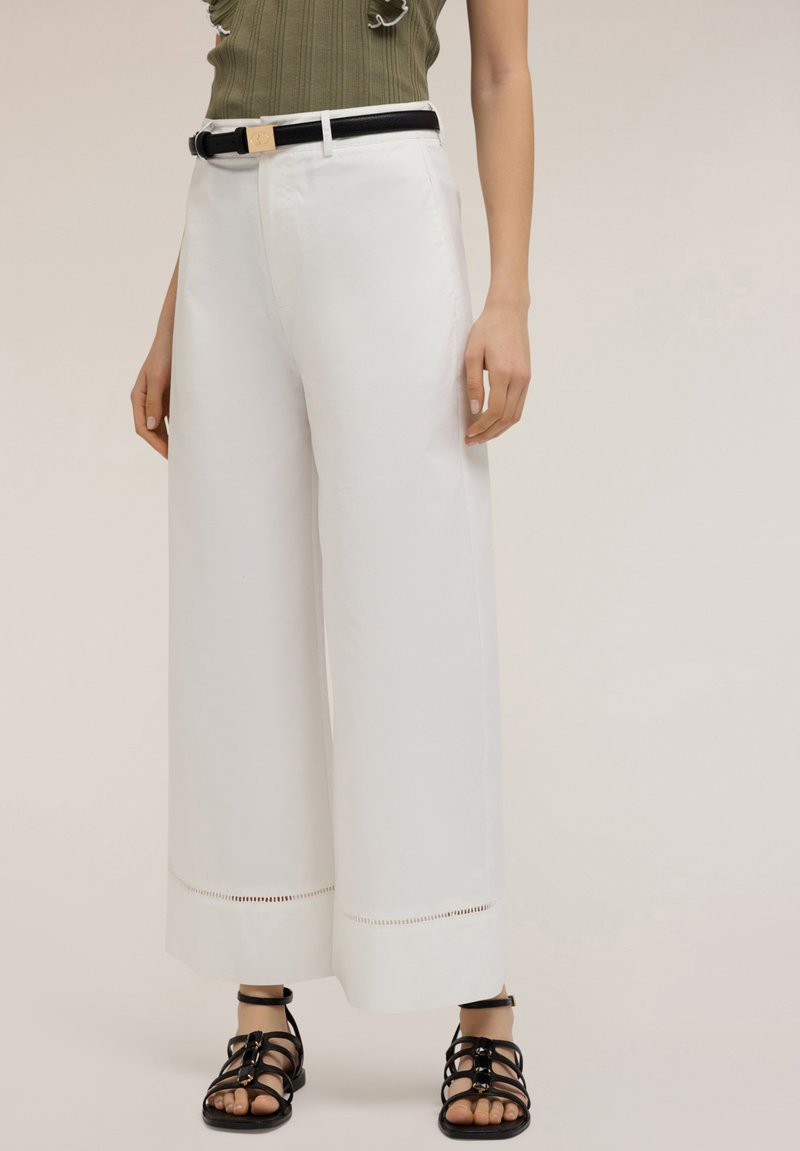 White wide-leg trousers feature a smooth texture with cut-out detailing at the hem. Paired with a black belt and strappy black sandals.