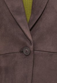 Bruine faux suède blazer met een notch revers, een enkele knoopsluiting en gestikte details, gelaagd over een groene gebreide top.