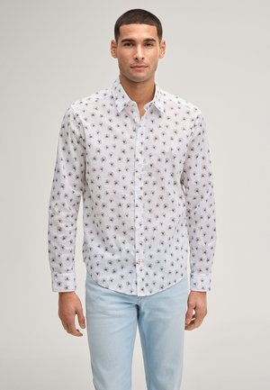 Uomo che indossa una camicia bianca a maniche lunghe con un piccolo motivo floreale scuro e jeans azzurri, in piedi davanti a uno sfondo semplice.
