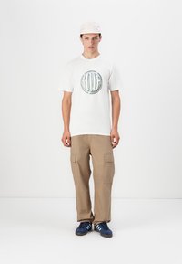 Hvid t-shirt med cirkulært grønt grafisk print, beige cargo-bukser med sidelommer og blå sneakers med tre hvide striber. Lys kasket.