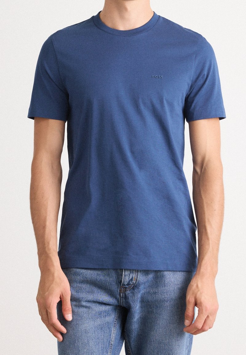 Homme portant un t-shirt bleu uni à manches courtes avec un petit logo "BOSS" sur la poitrine et un jean bleu clair sur un fond uni.