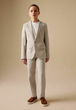 Boy standing, wearing a beige suit with a white shirt and brown slip-on shoes, against a plain beige background.