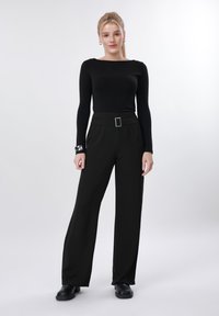 Ξανθιά γυναίκα στέκεται μπροστά σε λευκό φόντο, φορώντας μαύρο μακρυμάνικο τοπ, μαύρο παντελόνι ψηλόμεσο και φαρδύ με ζώνη και μαύρα παπούτσια.