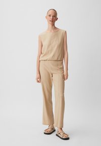 Beige ärmelloses Top, kombiniert mit passenden weit geschnittenen Hosen, beide aus strukturiertem Stoff gefertigt. Das Model trägt minimalistische Sandalen mit Riemendetails.