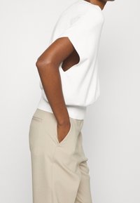 Bílý krátký rukáv top s otevřenými bočními částmi, s volným střihem a žebrovaným lemem. V kombinaci se světle béžovými kalhotami s bočními kapsami.