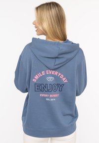 Blauer Kapuzenpullover mit einer frontalen Kängurutasche, verziert mit buntem Text: "LÄCHEL JEDEN TAG", "GENIEßEN" und "GRÜNDUNG 2014" in kontrastierender Stickerei.