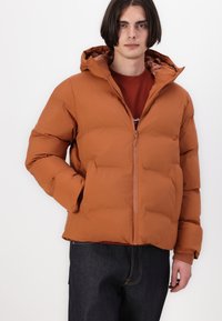 Brun dunjakke med hætte, med en frontlynlås, elastiske manchetter og en tekstureret overflade. Båret over en rød skjorte og mørke jeans.